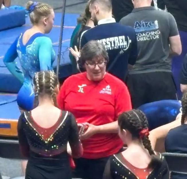 Woman in red shirt speaking with two girls in black leotards with braided hair.