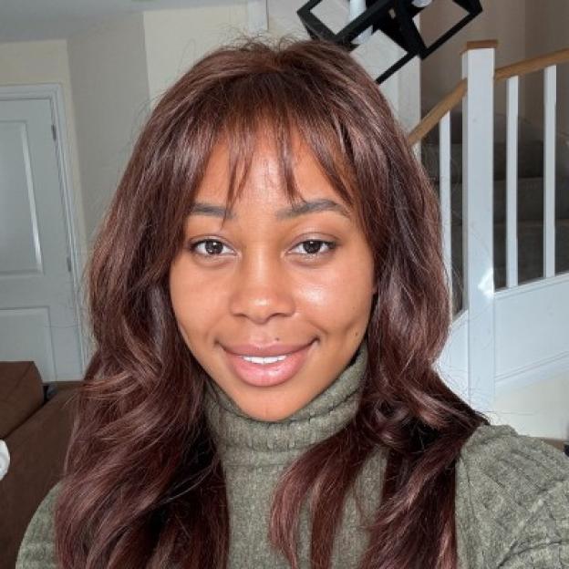 Headshot of Hadiza Atunse, a woman with long brown hair and bangs, smiling while wearing a green turtleneck.