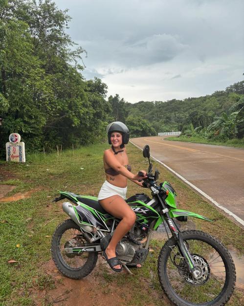 A young woman wearing a helmet and a bikini top sits on a green dirt bike next to a rural road, with lush green hills in the background.