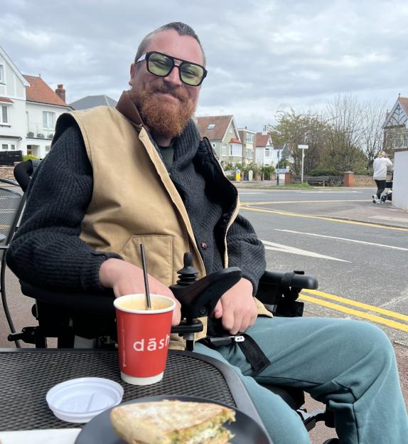 Liam Jefferies, 34, in his wheelchair, seated at an outdoor cafe with a panini and a drink on the table.