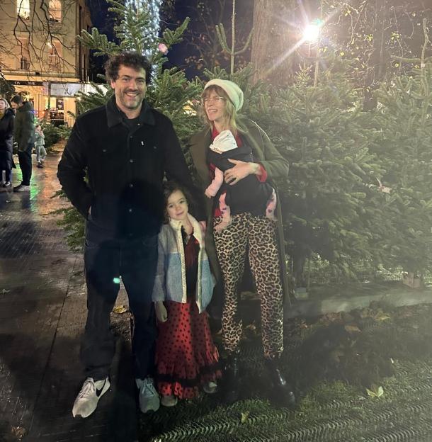 A family of four, including a baby in a carrier, standing in front of Christmas trees at night.