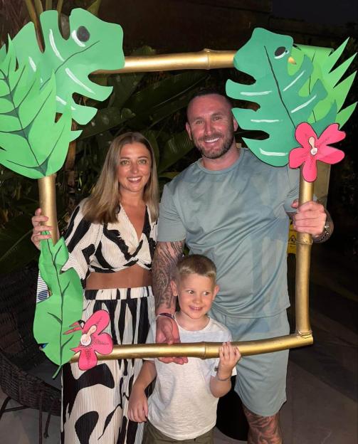 James Bainbridge with partner Jessica Hill and son Alfie posing in a photo frame with tropical leaf decorations.