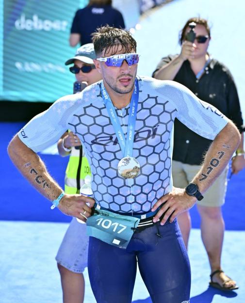 A male triathlete wearing a light blue and dark blue suit, sunglasses, and a medal around his neck, with bib number 1017.