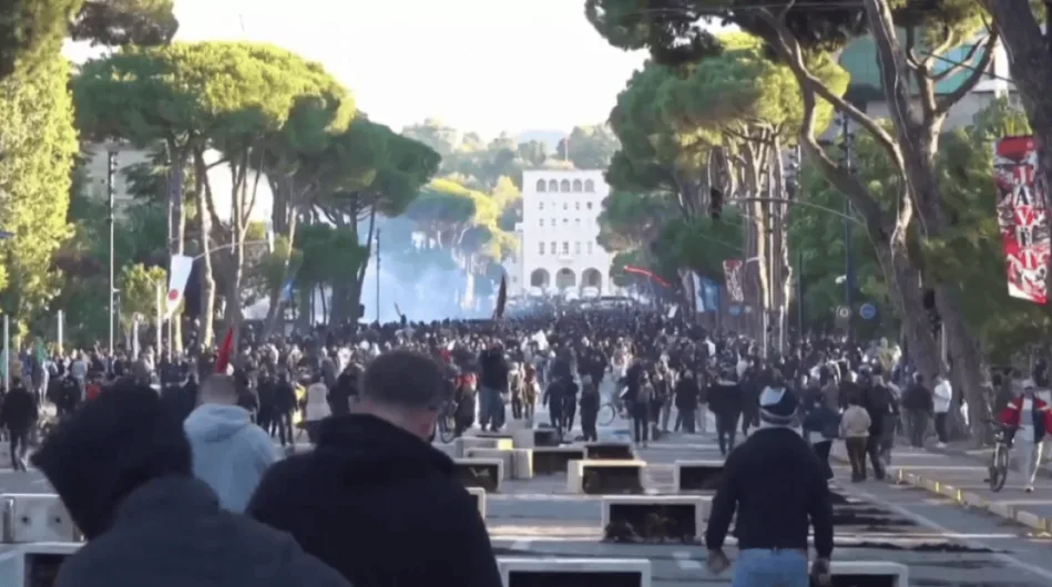 A large crowd of protesters fills a tree-lined boulevard in Tirana, Albania, moving toward a white building in the background.