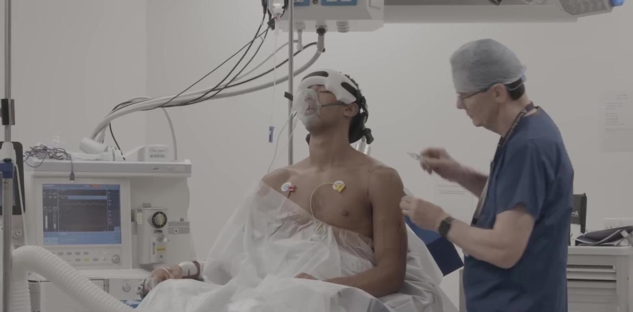 Jude Bellingham in a hospital gown, attached to medical equipment, while a doctor prepares for his shoulder surgery.