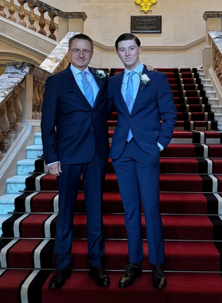 Two men in blue suits stand on a grand red carpeted staircase.
