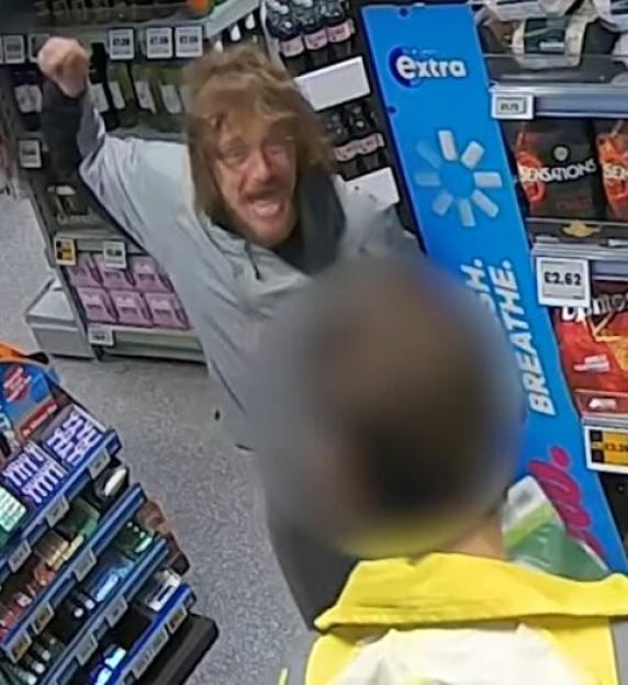 A man with disheveled hair and a mustache, wearing a gray jacket, shouting with an aggressive expression in a store aisle.