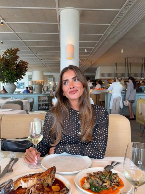 A woman in a black and white polka dot shirt with white wine and food at a restaurant.