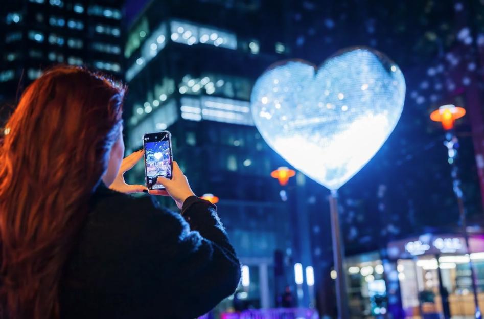Woman photographing a large, glowing heart light installation with her phone at night.