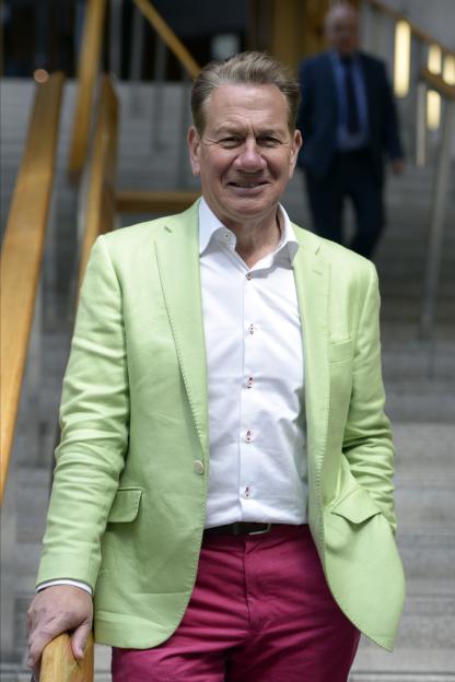 Broadcaster Michael Portillo in a bright green jacket and pink trousers, standing on steps in the Scottish Parliament.