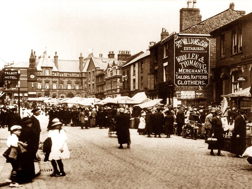 Ormskirk Market Day in the early 1900s, featuring a large crowd gathered on a cobbled street lined with market stalls and buildings, including the Talbot Hotel and a "Woollen & Trimming Merchants" shop.