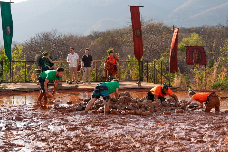 Four contestants from "I'm A Celebrity... Get Me Out Of Here! South Africa" in a muddy challenge, with two hosts observing.