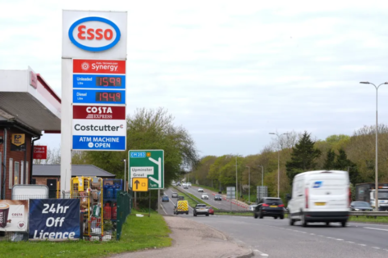 An Esso gas station sign showing prices for unleaded (159.9) and diesel (194.9), alongside Costa Express and Costcutter branding, with a road leading to Upminster Great and the M25 in the background.