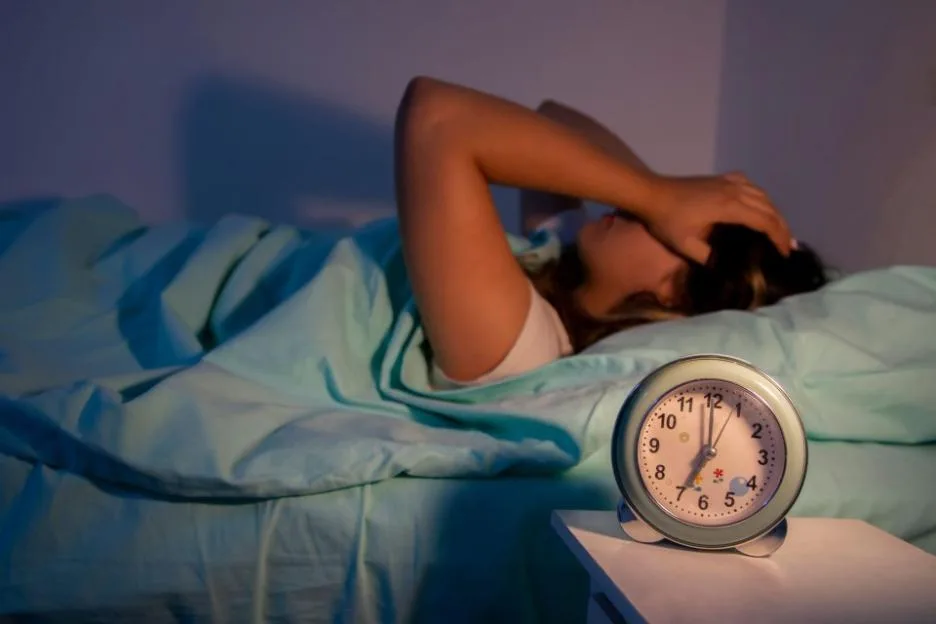 A teenage girl lying in bed next to an alarm clock struggling to wake up.