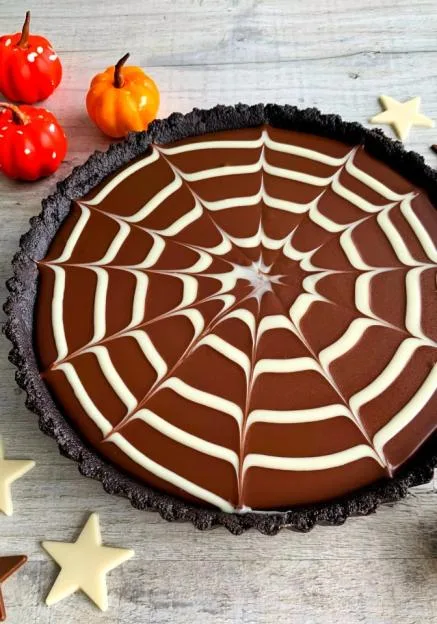 A chocolate tart with a white chocolate spider web design on top, accompanied by small decorative pumpkins.