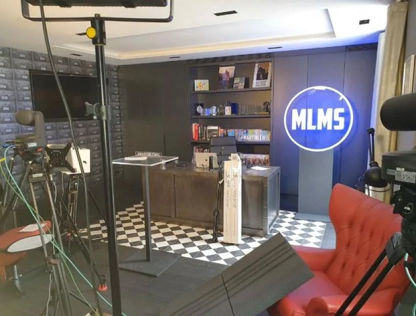 A room set up as a home TV studio, with tripods, cameras, and a glowing "MLMS" sign.