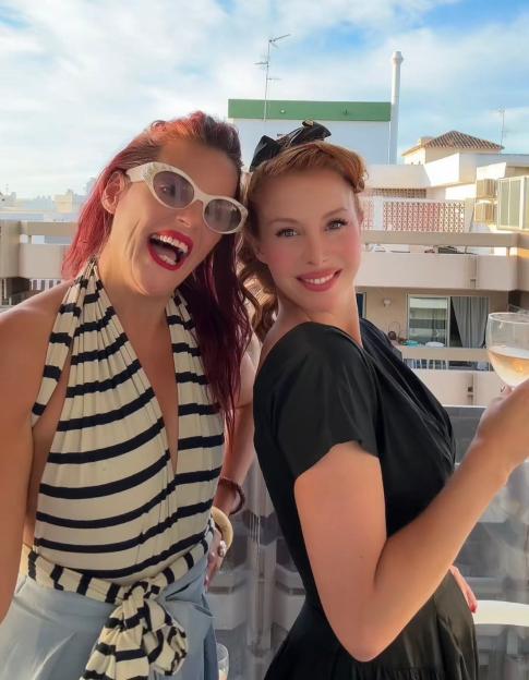 Two women on a balcony, one with red hair and white cat-eye sunglasses smiling, and the other with auburn hair holding a glass of wine, against a backdrop of buildings and a partly cloudy sky.
