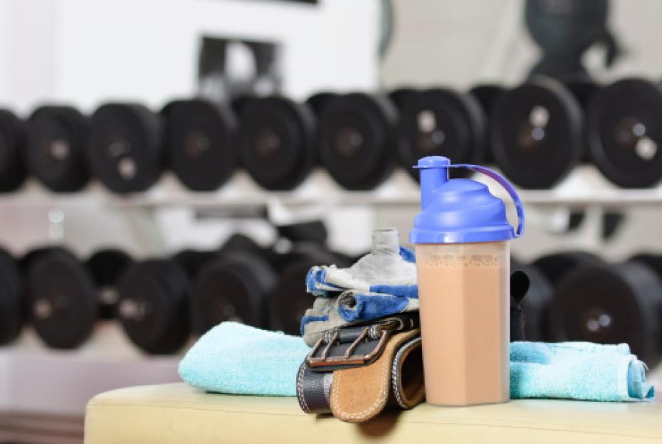 Protein shake, towel, weightlifting gloves, and belt on a bench in a gym with dumbbells in the background.