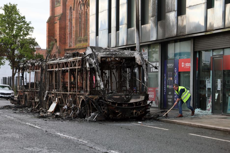 A man sweeping glass from the street next to a burnt-out bus and a damaged storefront in East Belfast.