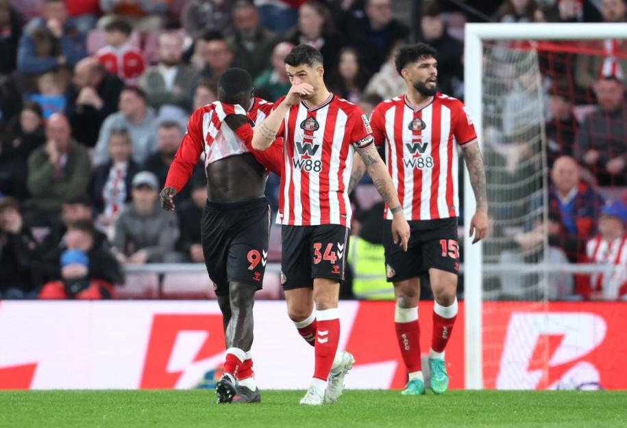 Three football players in red and white striped jerseys with black shorts walk on a green field at the Stadium of Light.