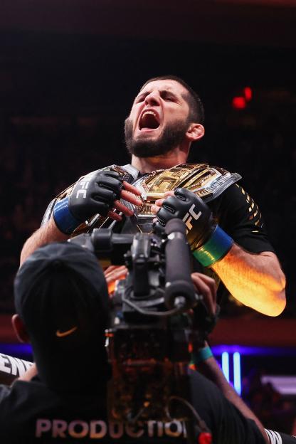 Islam Makhachev celebrates his welterweight championship victory while wearing blue gloves and a gold belt.
