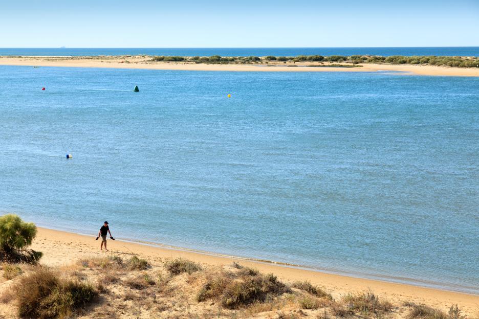 El Rompido beach in Costa de la Luz, Andalusia, Spain, with a person walking on the sandy shore next to calm blue waters and an island in the distance.
