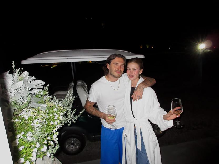 Brooklyn Beckham with his arm around Nicola Peltz, both holding wine glasses, in front of a golf cart at night.