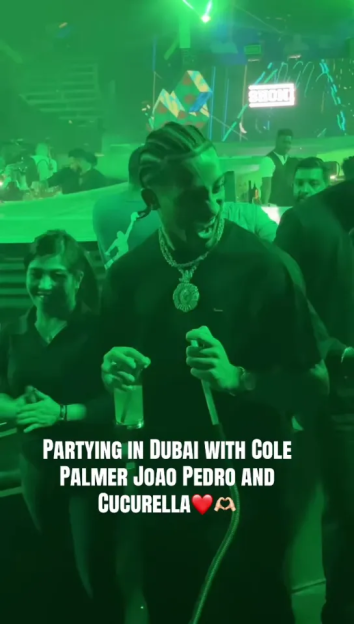 A man wearing a heavy gold chain and black t-shirt smiles at a party.