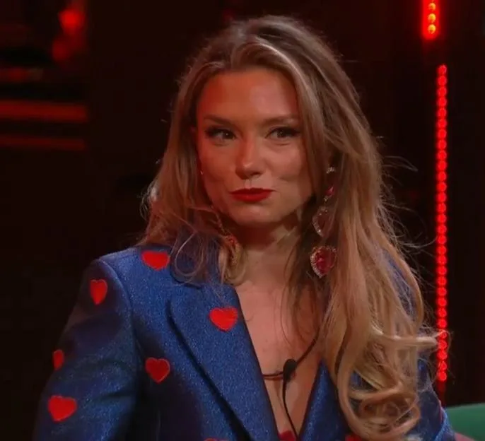 A woman with blonde hair, red lipstick, and heart earrings, wearing a blue jacket covered in red hearts.