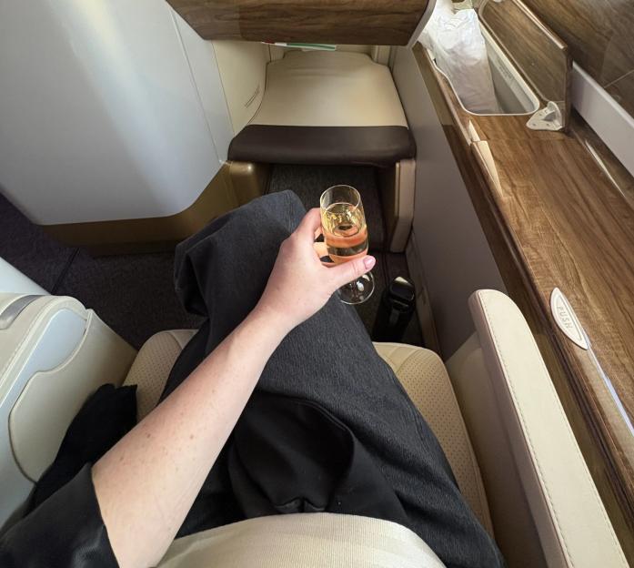 A person sitting in a luxurious airplane seat, holding a glass of champagne, with an inflight entertainment screen displaying "P.A. in progress" and "Travel Kit Essentials".