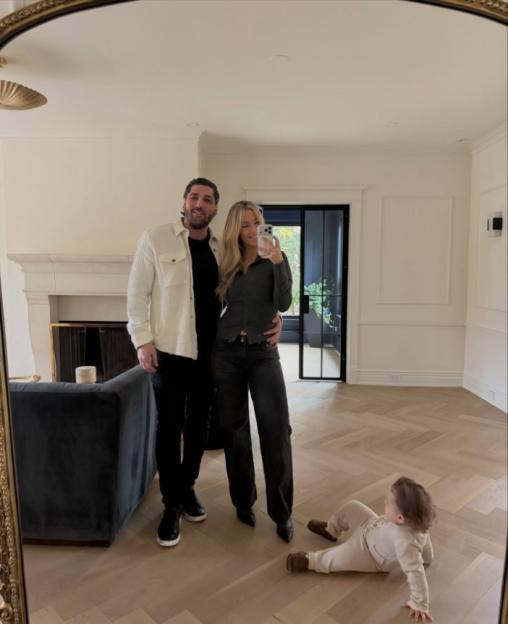 A couple and a baby in a mirror selfie inside a large, unfurnished room with light wood floors.