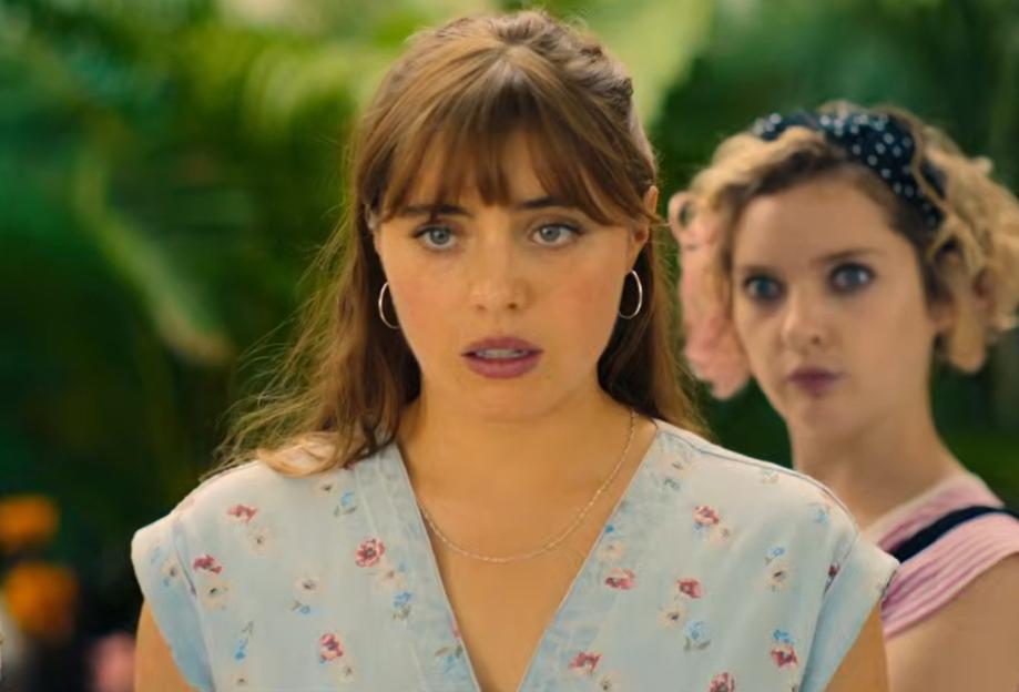 A still from the TV series "Rivals" shows a young woman with brown hair, blue eyes, and bangs, wearing a light blue floral dress, looking shocked. Another woman is blurred in the background.