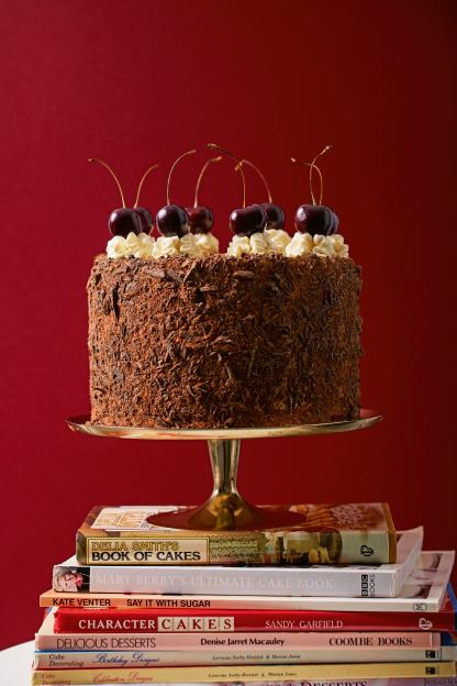 A black forest cake covered in chocolate shavings, topped with whipped cream and cherries, rests on a gold cake stand which sits on a stack of cookbooks.
