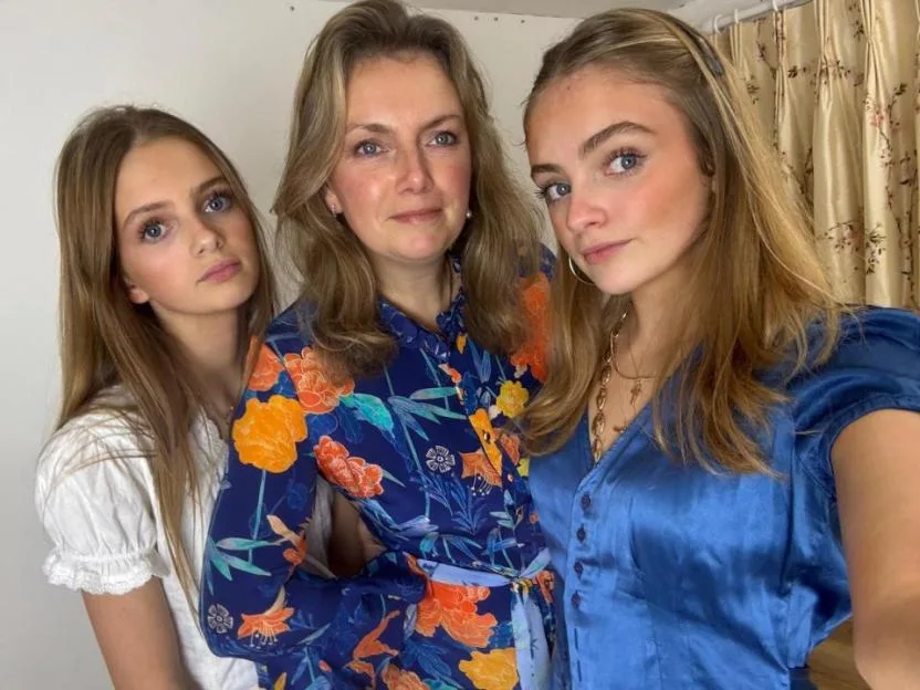 A mother and two daughters standing together, all with light brown hair and blue eyes.