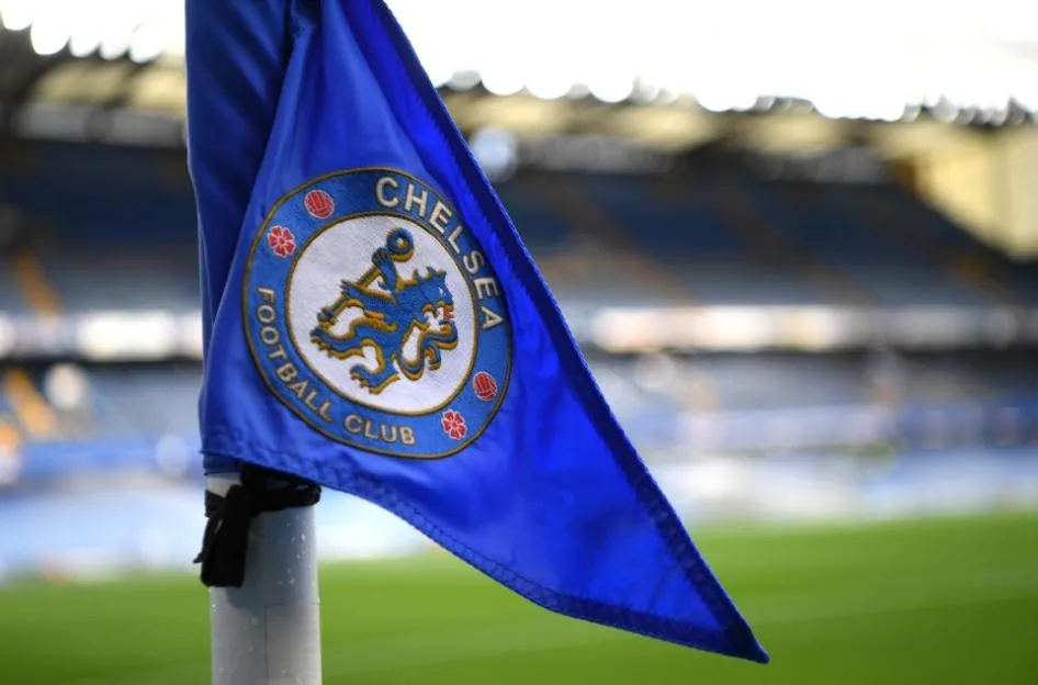 A blue corner flag featuring the Chelsea Football Club crest, with the Stamford Bridge stadium blurred in the background.