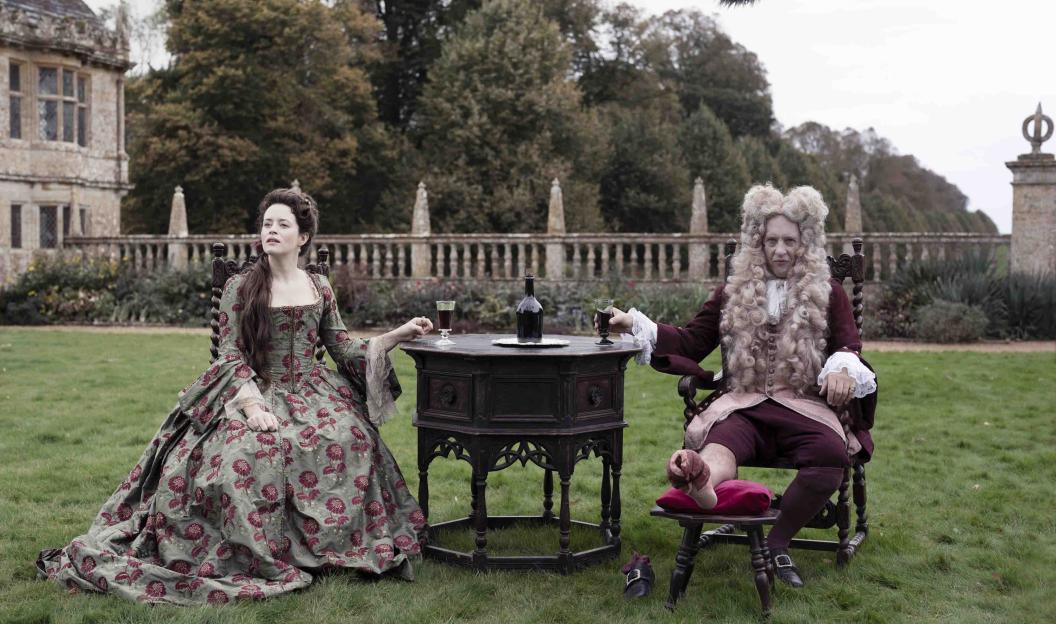 A woman in an elaborate dress and a man in a wig and period attire sit at an outdoor table with drinks, with an old building in the background.