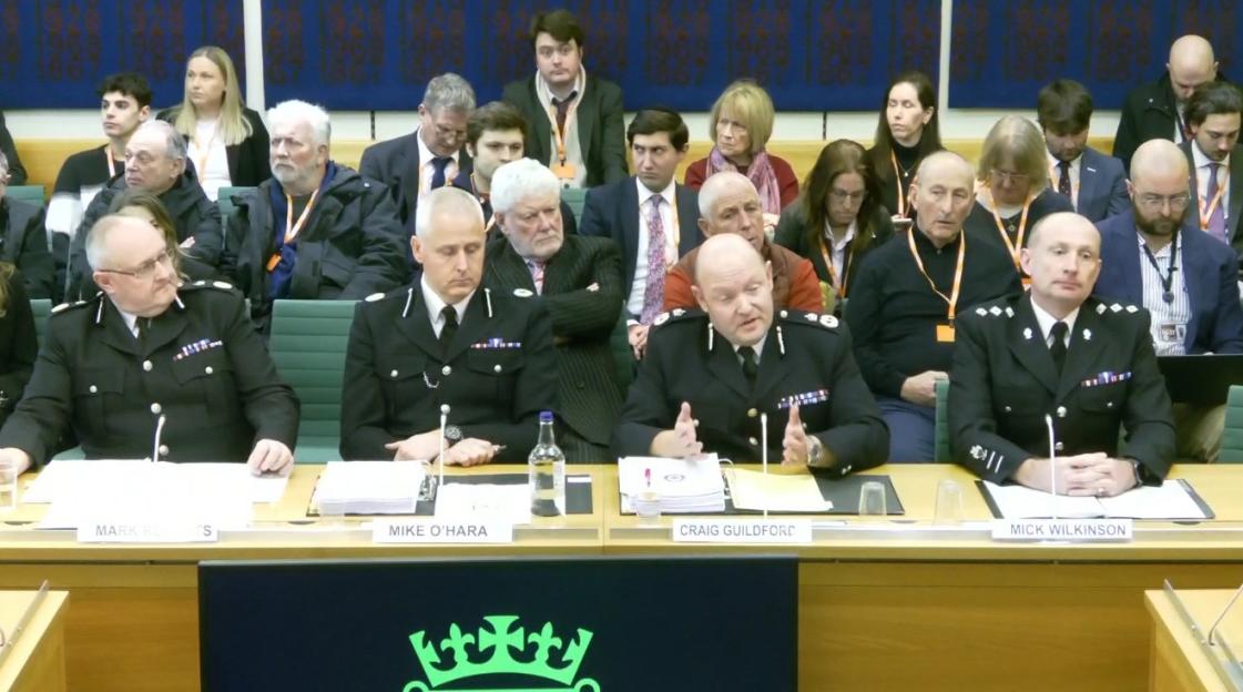 Four uniformed police officers, Mark Roberts, Mike O'Hara, Craig Guildford, and Mick Wilkinson, sit at a table facing a group of people, with Craig Guildford speaking during a hearing.