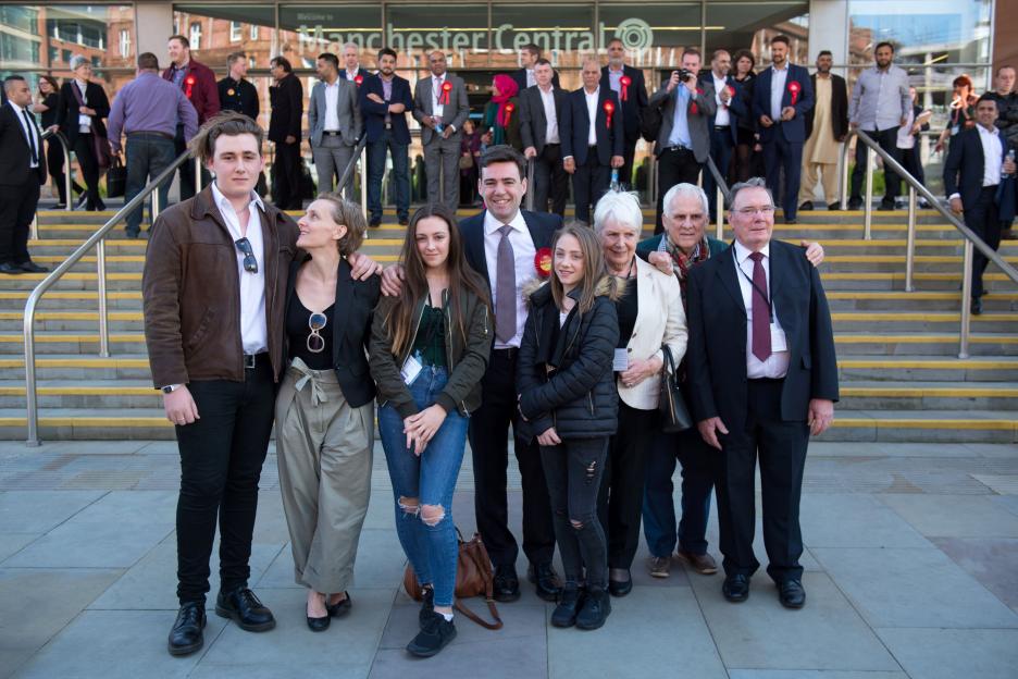 A group of people, some wearing "Vote" rosettes, standing on steps in front of a building with "Manchester Central" written on it.
