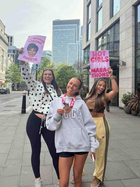 Megan Moore and Lauren Wood smiling and holding signs, one reading "HOT GIRLS RUN MARATHONS" and another with Kris Jenner's face.