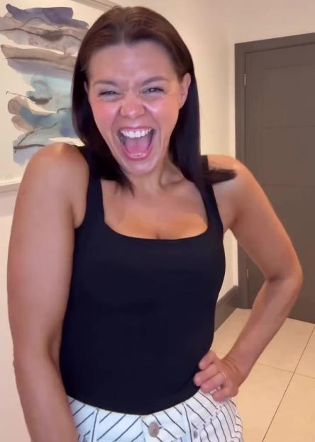 A woman in a black tank top and striped shorts laughs with her mouth wide open.
