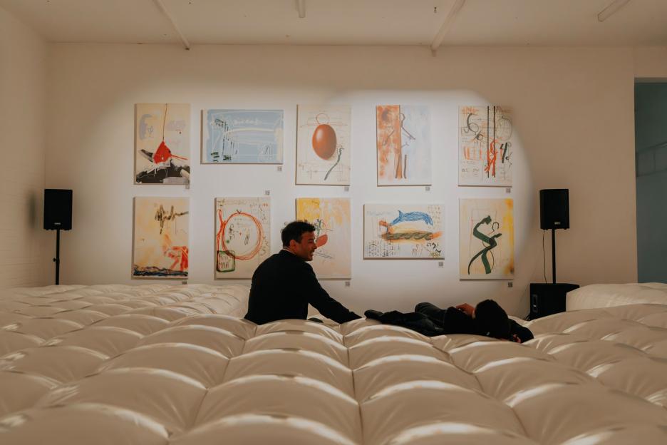 Two men relaxing on an inflatable mattress in a gallery filled with abstract paintings.