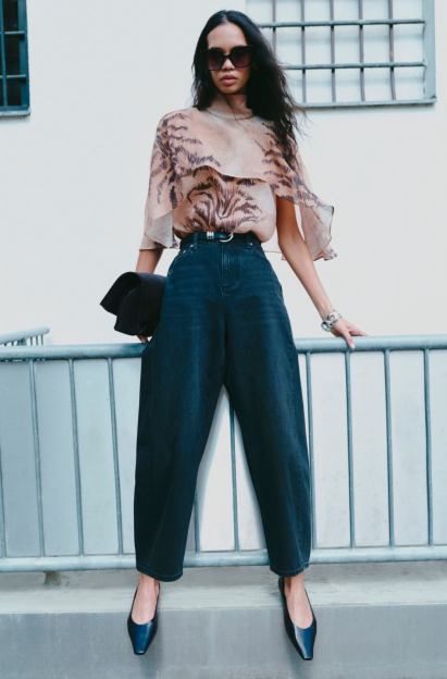 A model in a sheer ruffled blouse with a dark animal print, wide dark jeans, and black flats, standing behind a railing.