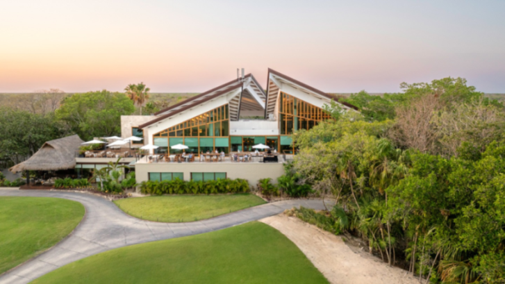 An aerial view of a large modern building with a restaurant patio and a thatched-roof building, surrounded by green golf course fairways and lush trees.