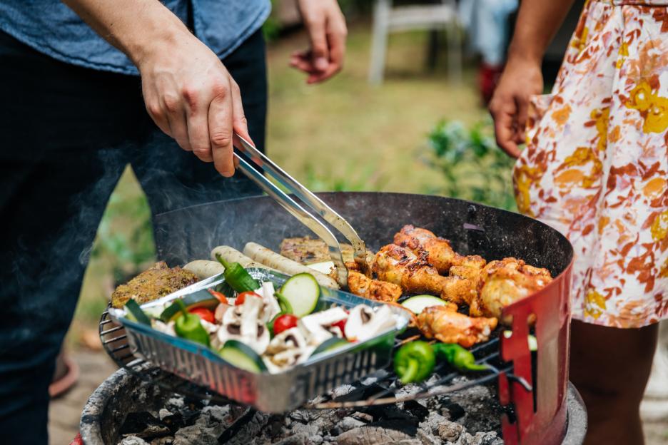 A person using tongs to cook various foods on a barbecue grill.