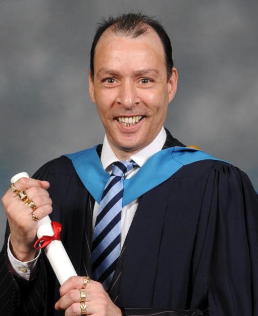 Alan Paul in a graduation gown, holding a diploma and wearing multiple rings.