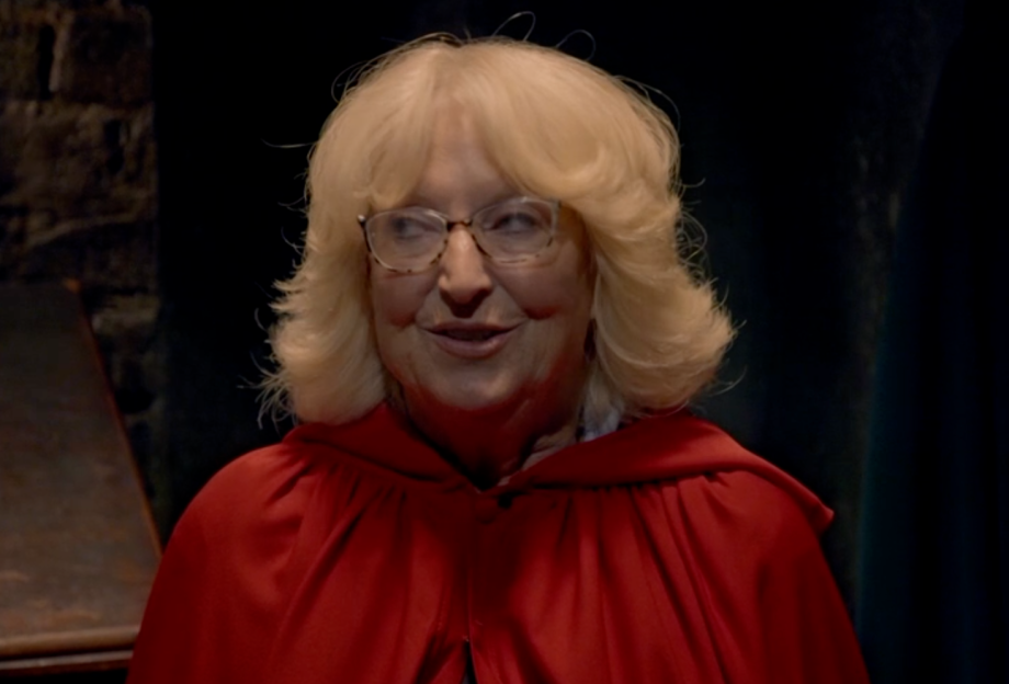 An older woman with blonde hair and glasses wearing a red cape, smiling.