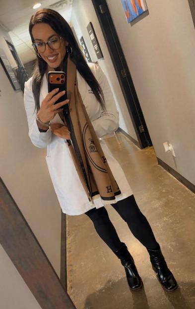 Crystal Janke, wearing a white lab coat and brown scarf, takes a selfie in a hallway.