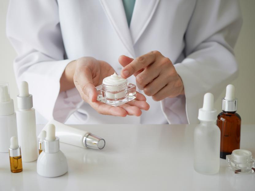 A person in a lab coat testing cream from a jar with their fingers.