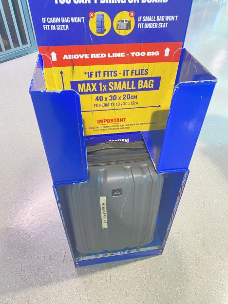 A gray suitcase barely fits into an airline baggage sizer, with a sign warning about oversized bags.