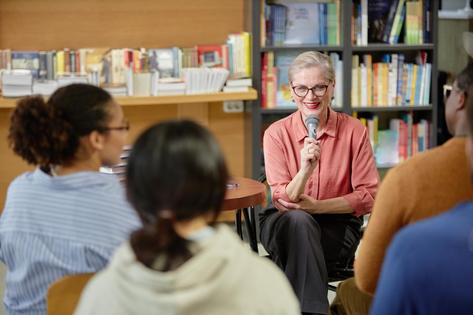 Senior Caucasian woman smiling and speaking into a microphone to a diverse audience in a library.